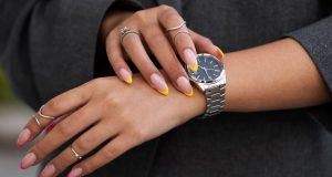 Stylish hands adorned with rings showcase a silver watch and vibrant yellow-tipped nails, highlighting modern fashion trends.