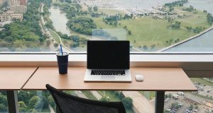 A modern workspace with a laptop and drink, overlooking a green park and marina from a high-rise window
