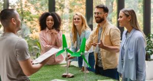 Team of young professionals discussing a miniature wind-turbine model in a sunlit office, showcasing corporate sustainability.