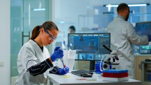 Scientist pipetting samples in high-tech biotech lab; microscope and DNA-analysis monitor underscore cutting-edge drug discovery research.