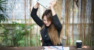 Frustrated woman raising laptop in anger over impulse spending and financial stress at outdoor workspace.